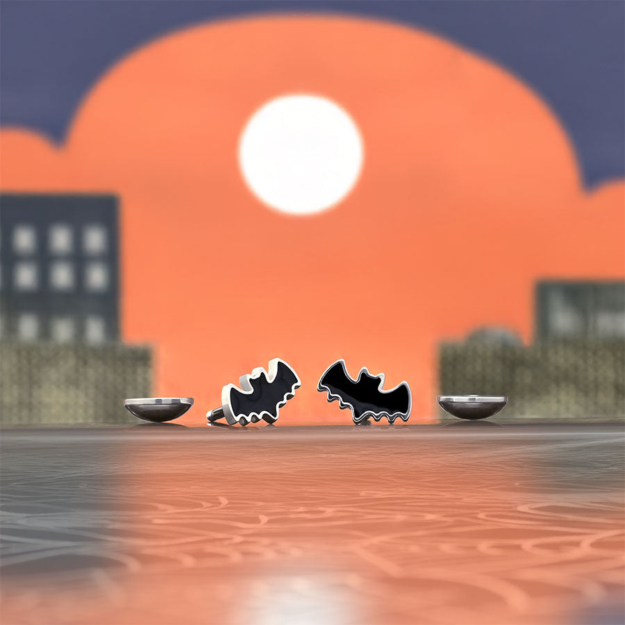 Two black bat-shaped earrings on a webbed surface with a cityscape and sunset in the background.
