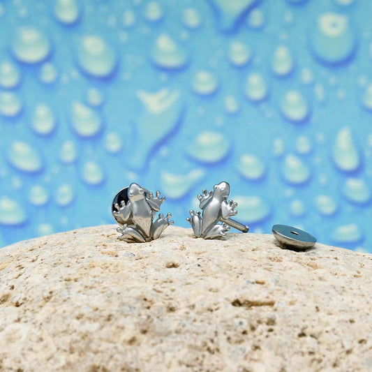 A pair of frog stud earrings displayed on a light beige stone, with a blue water-droplet-patterned background.