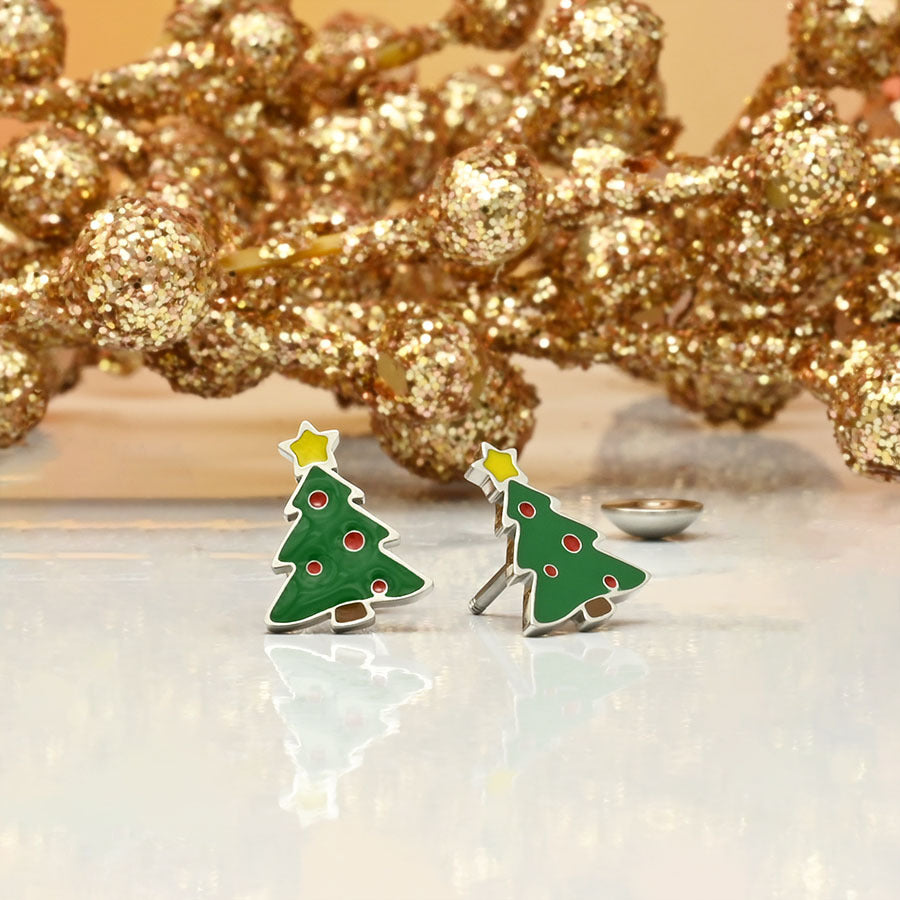 Green Christmas tree earrings with red dots and gold accents on a reflective surface with decorative gold branches in the background.