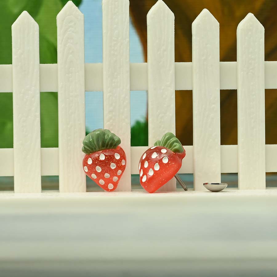 Two strawberry-shaped homemade DIY ComfyEarrings on a white picket fence with a blurred background
