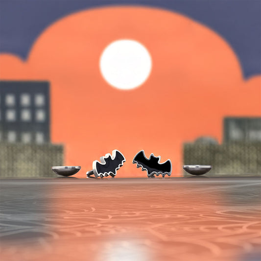 Two black bat-shaped earrings on a webbed surface with a cityscape and sunset in the background.