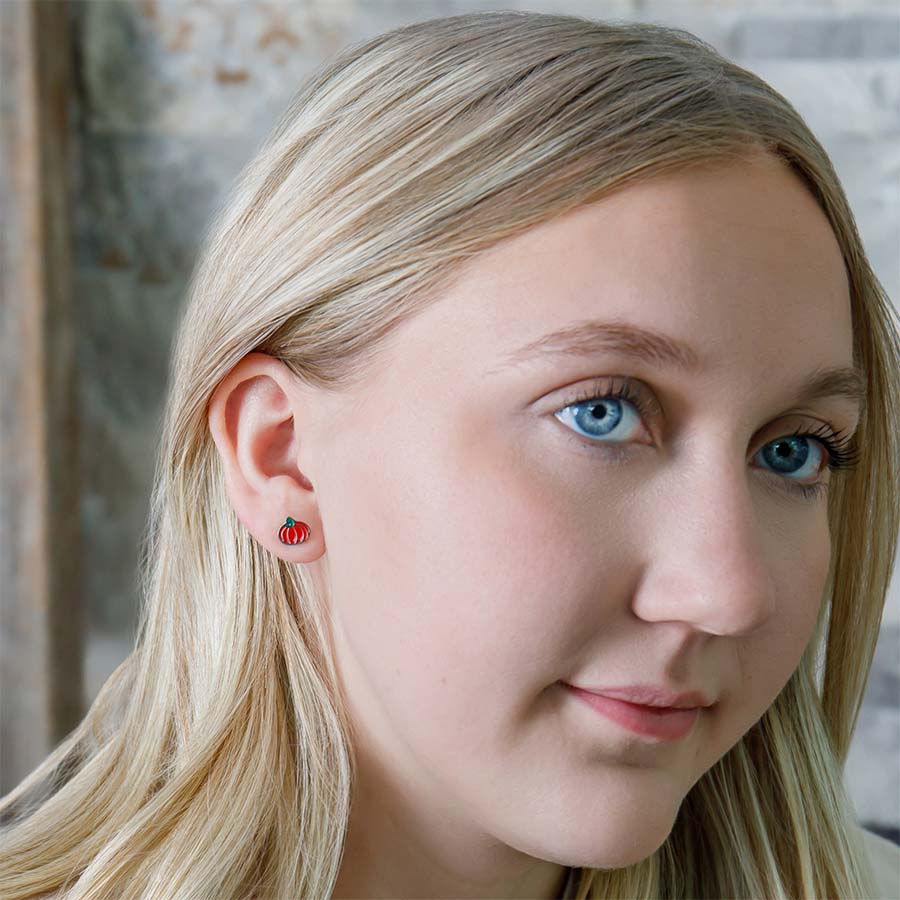 Close-up of a person wearing with small orange pumpkin earrings, against a neutral background.