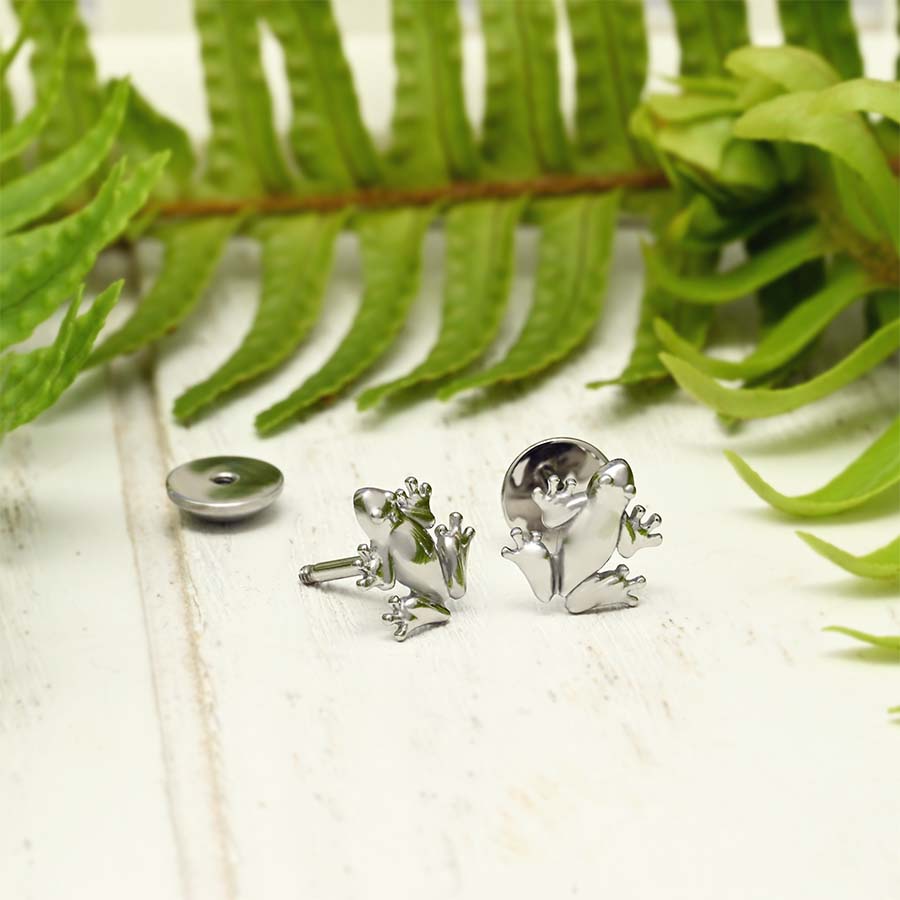A pair of frog stud earrings displayed on a white wooden surface with green fern leaves in the background. One earring is shown front-facing, revealing the detailed frog body and head, while the other is turned to show the back with its screw-on backing.