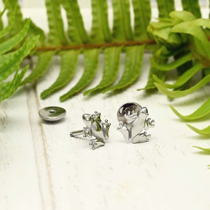 A pair of frog stud earrings displayed on a white wooden surface with green fern leaves in the background. One earring is shown front-facing, revealing the detailed frog body and head, while the other is turned to show the back with its screw-on backing.