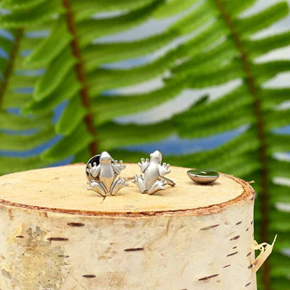 A pair of frog-shaped stud earrings displayed on a birch log stump, blurred green fern leaves in the background.