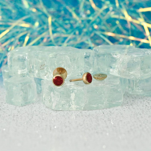Ruby Red Rose Gold ComfyEarrings sitting on clear glass chunks and blue sparkly background.