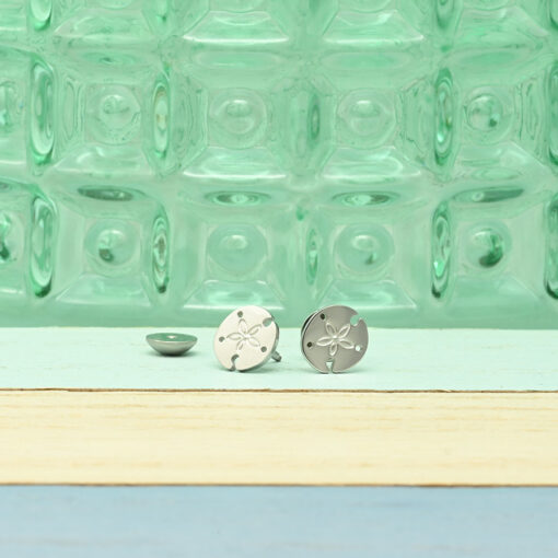 Stainless Sand Dollar ComfyEarrings sitting on a beach board in front of green patterned glass.